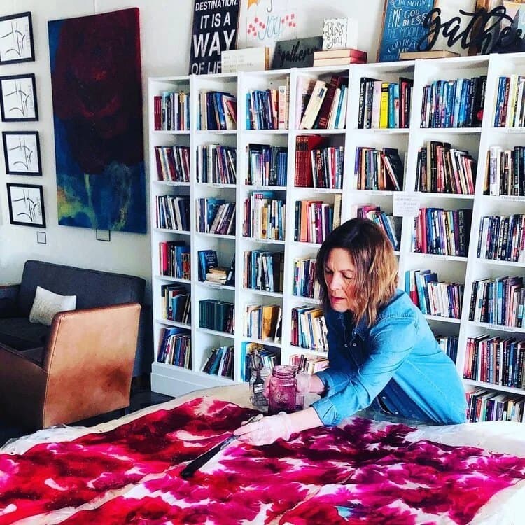 Barbie Hunt painting a crimson silk in her studio, shelves of books behind her